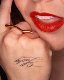 Amelia sculptural gold wave ring worn with black nails in a close hand-to-lips portrait.