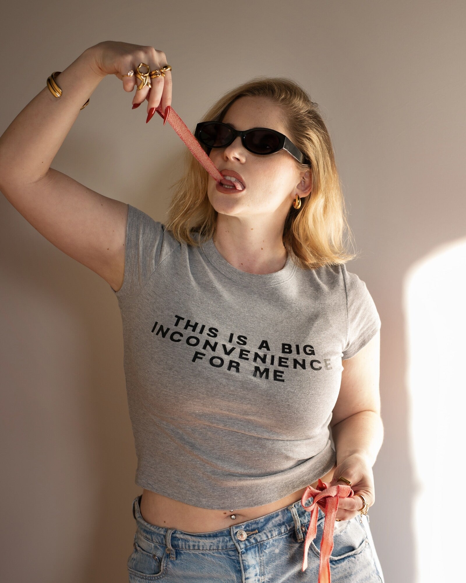 Model wearing the gray This Is a Big Inconvenience baby tee with sunglasses and red accessories.