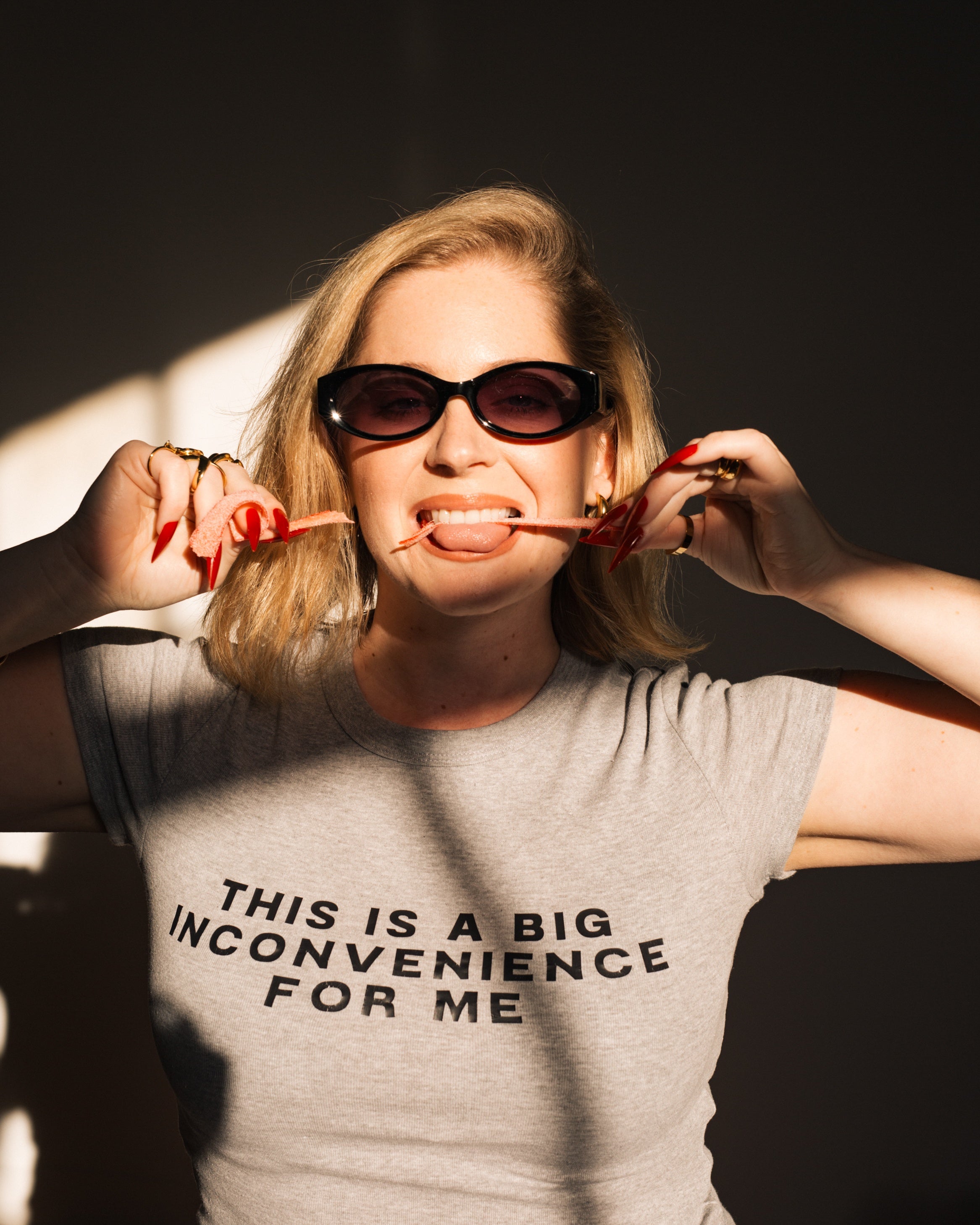 Model styling the gray Big Inconvenience baby tee with black sunglasses and red accessories.