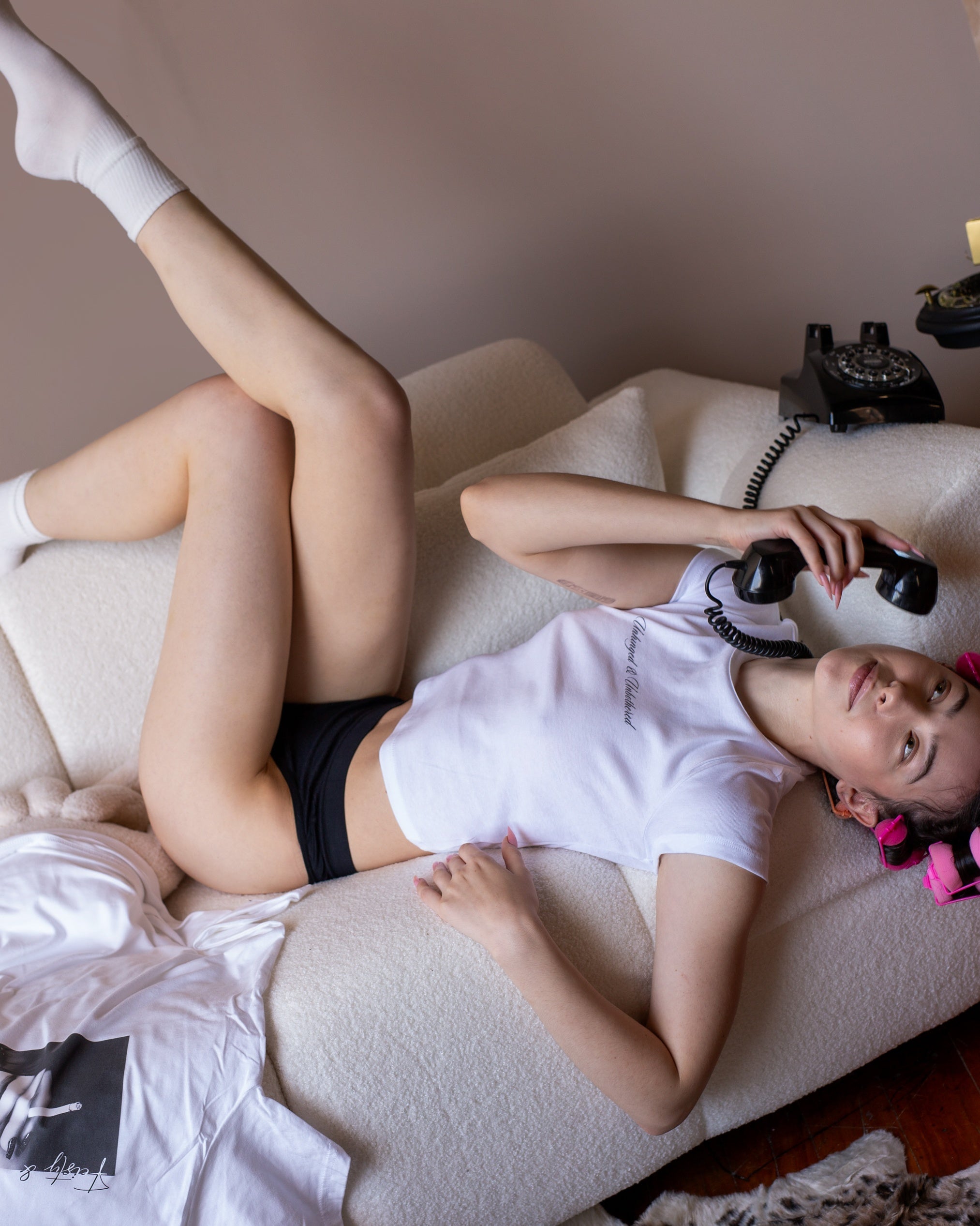 Model reclining in the white Unhinged and Unbothered baby tee with pink rollers and a black telephone.